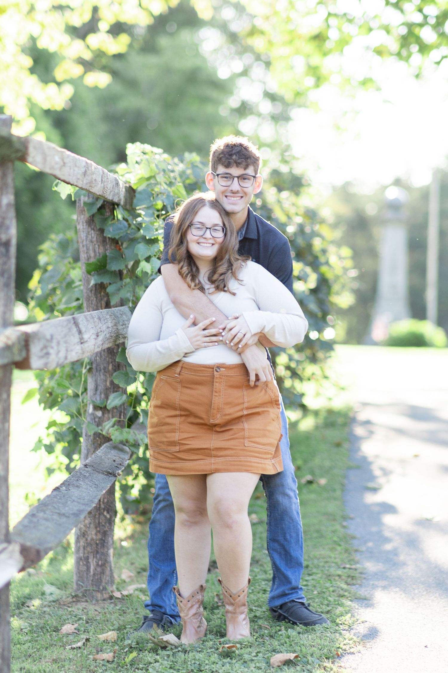 A couple poses outdoors by a rustic fence, woman in tan skirt and white top, man in dark polo embracing her from behind.
