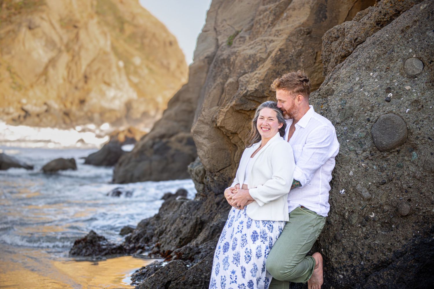 Couple embracing by rocky coastal cliffs on a sandy beach with ocean waves in the background.