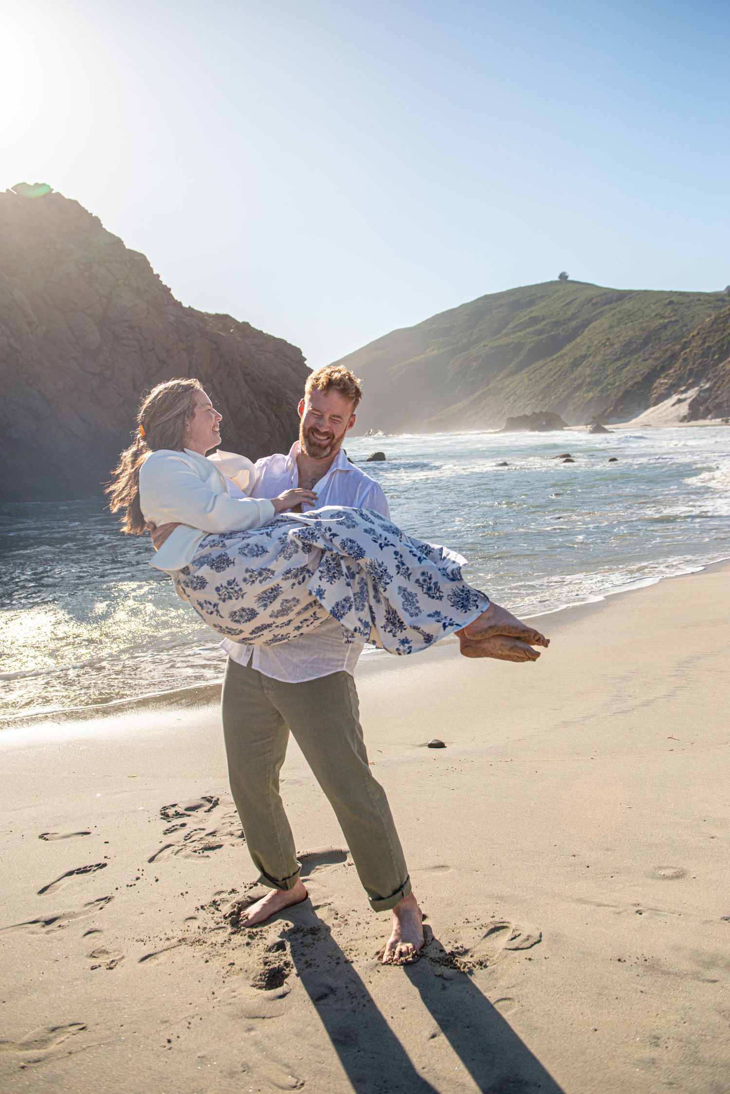 Man carrying woman in floral dress on sunny beach with dramatic cliffs and ocean waves in background.