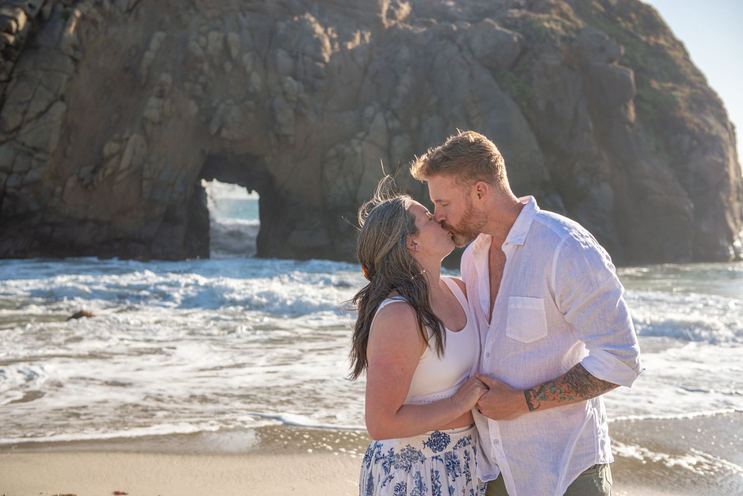 Couple sharing a romantic kiss on a rocky beach with ocean waves and a natural rock arch in the background.