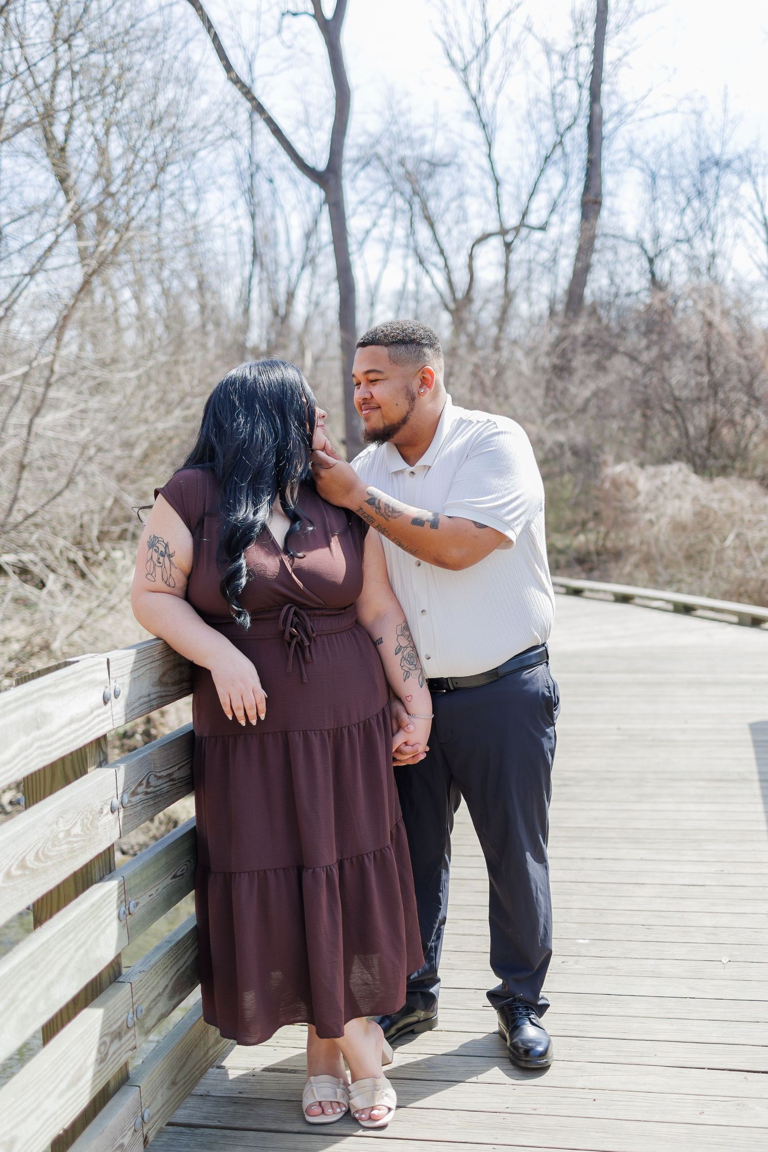 Couple sitting on grass, man hugging woman from behind, both wearing glasses, smiling, with tattoos visible outdoors.