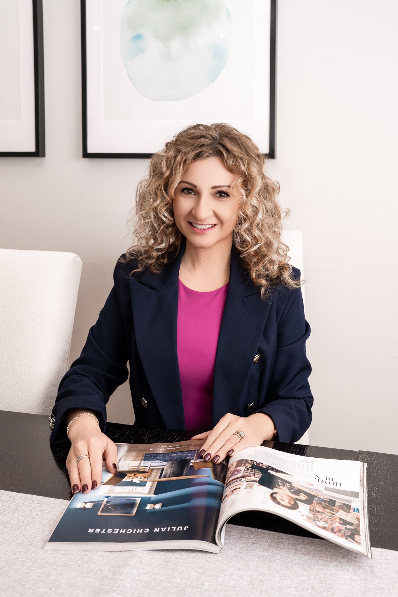 Smiling woman with curly blonde hair in navy blazer and pink top reviewing a Julian Chichester catalog at a desk.