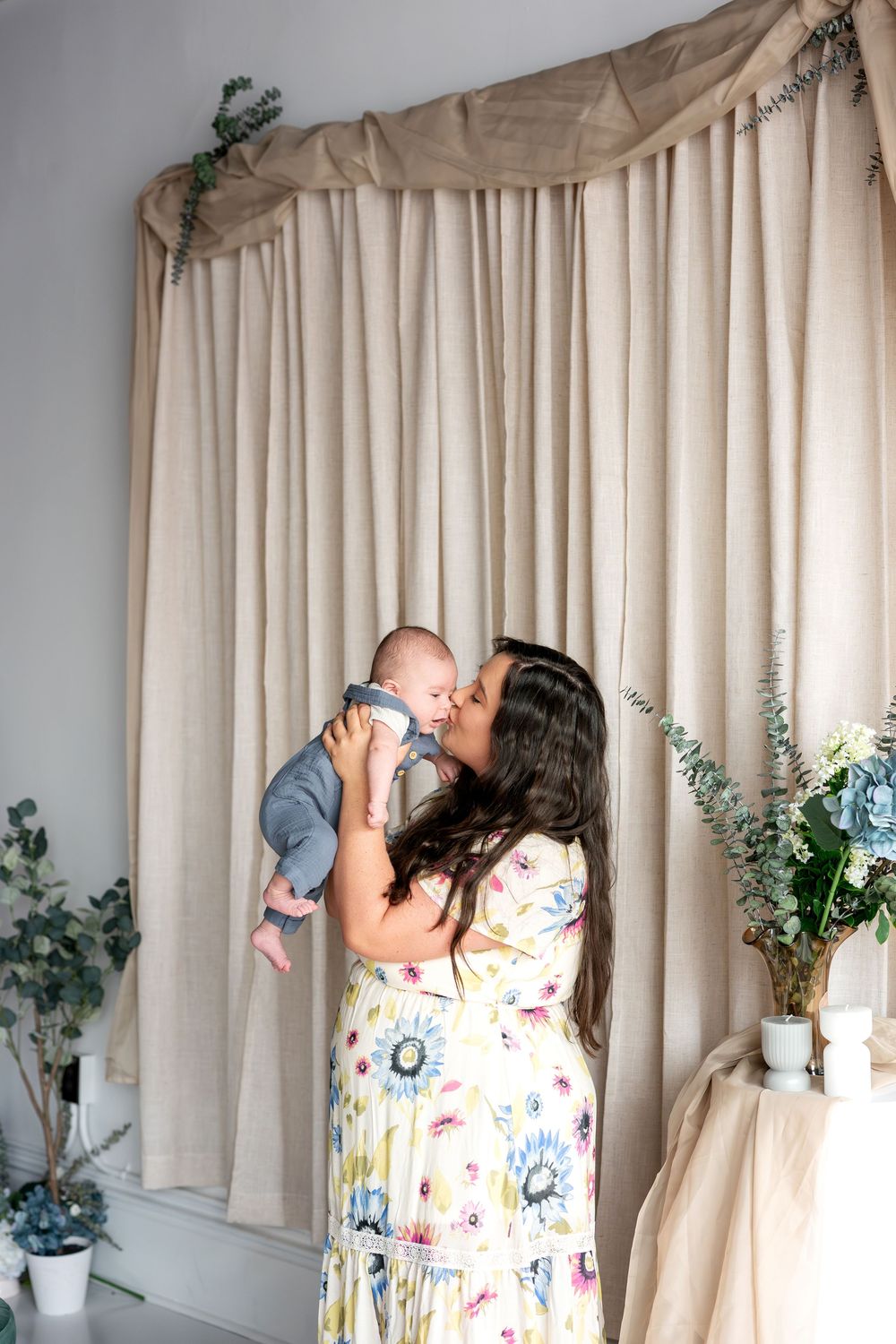 Mother in floral maxi dress kissing her infant baby lifted up near linen curtain backdrop.