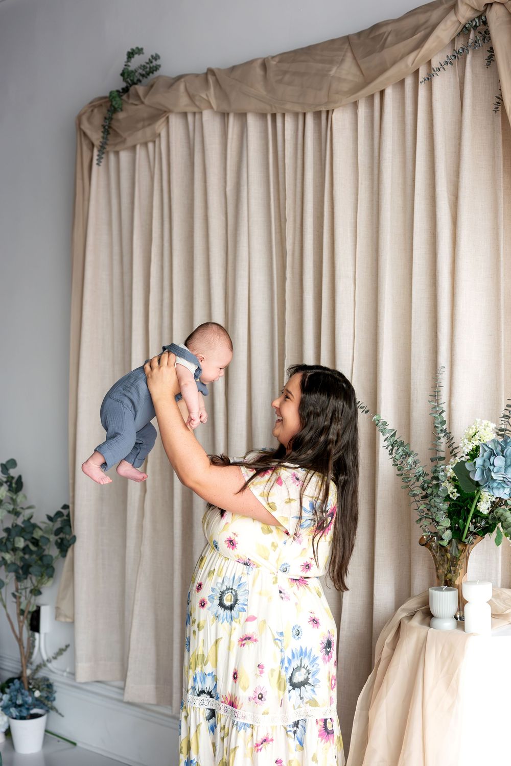 Joyful mom in floral dress lifting smiling baby high in air against neutral linen curtain backdrop.