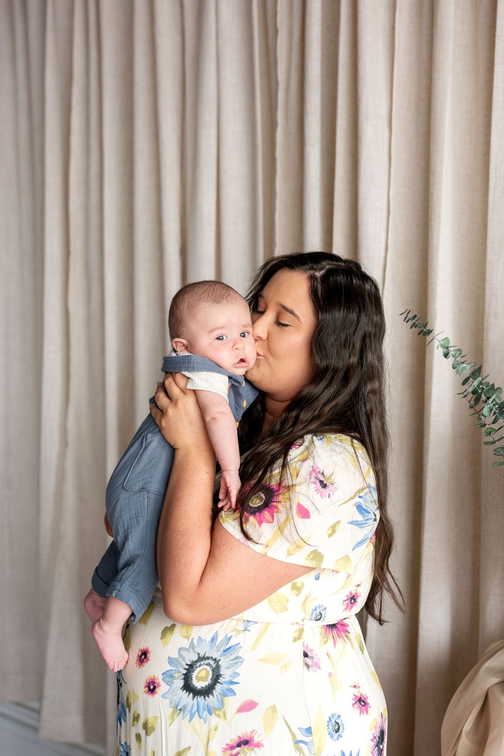 Mother in colorful floral dress gently kissing her baby boy in front of linen curtain backdrop.
