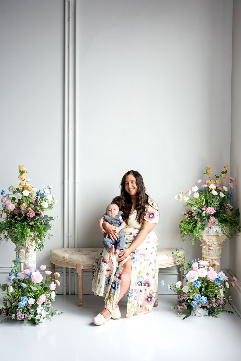 A smiling mother in a floral dress holds her newborn baby on a bench surrounded by colorful flower arrangements.