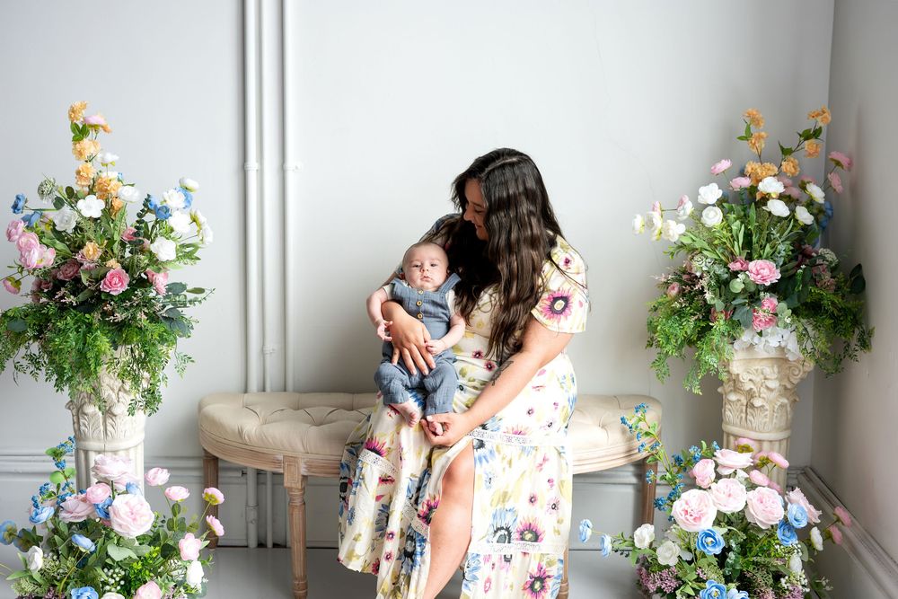A mother in a floral dress tenderly kisses her newborn baby while seated among lush flower displays.