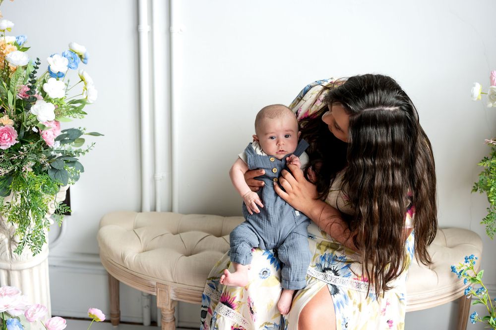 A mother in a floral dress gently nuzzles her smiling newborn baby surrounded by soft floral arrangements.
