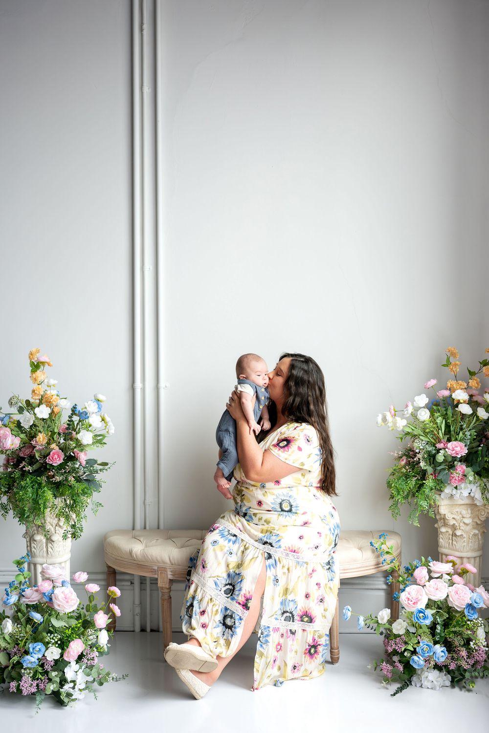 A mother in a floral maxi dress lifts and kisses her newborn baby surrounded by colorful floral displays.