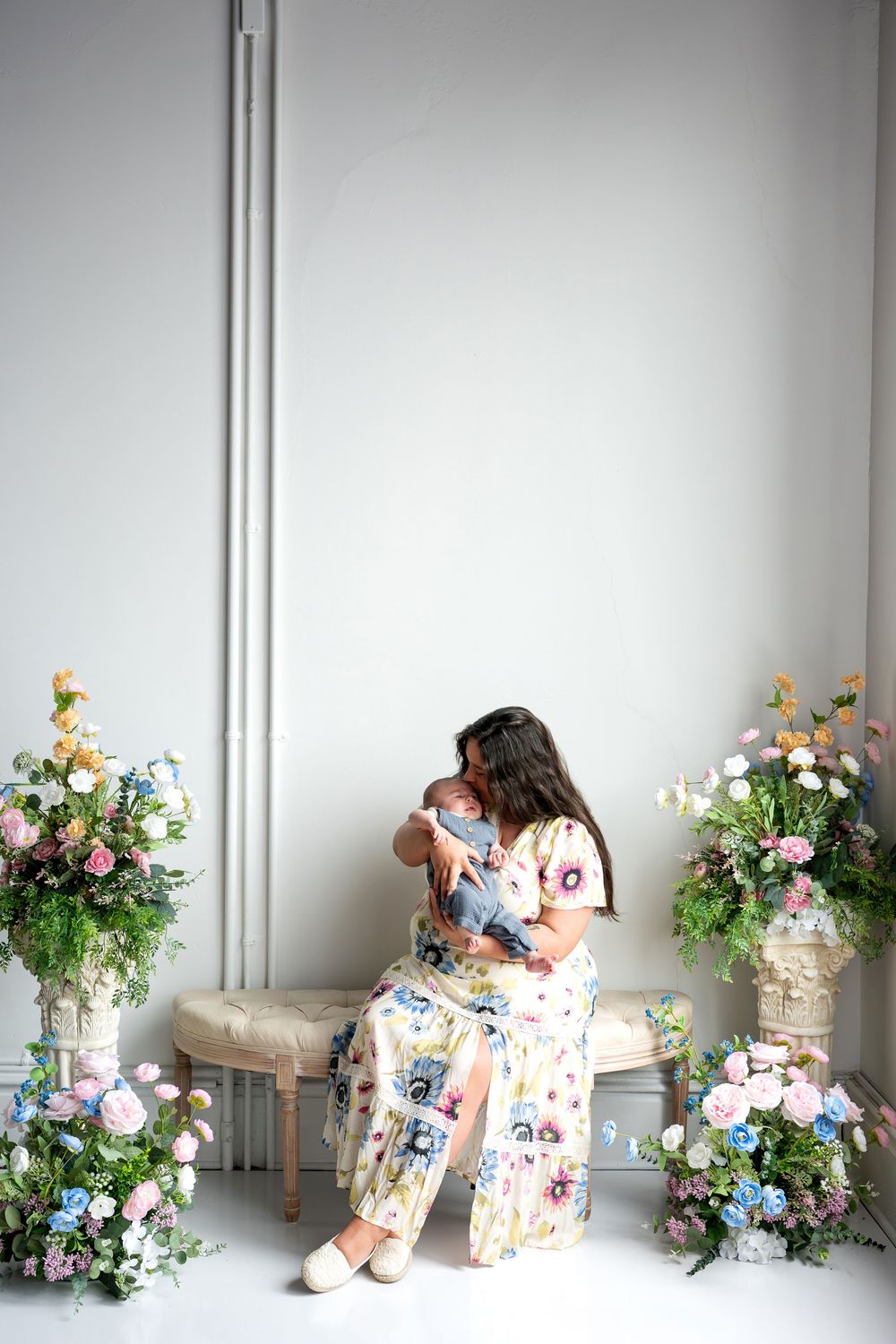 A mother in a floral dress cradles and kisses her sleeping newborn on a bench with flower arrangements.