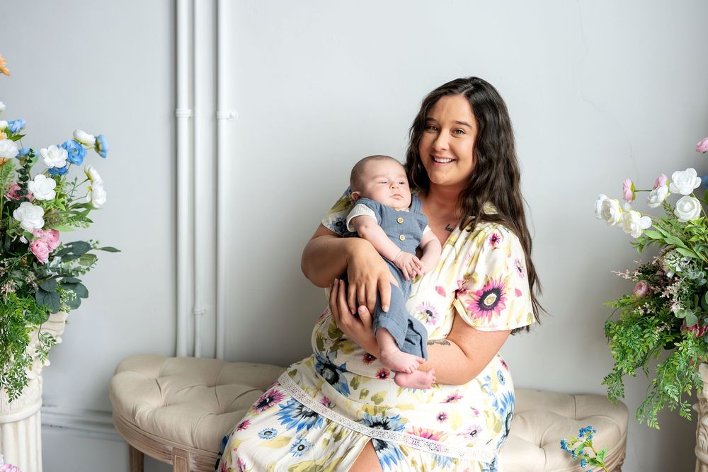 A smiling mother in a floral dress holds her newborn baby close on a bench with pastel flowers nearby.