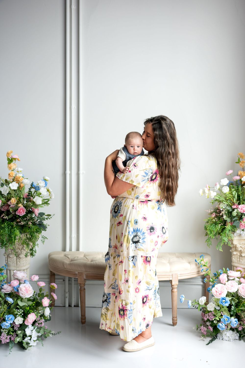 Mother in floral maxi dress kissing newborn baby surrounded by elegant floral arrangements indoors.
