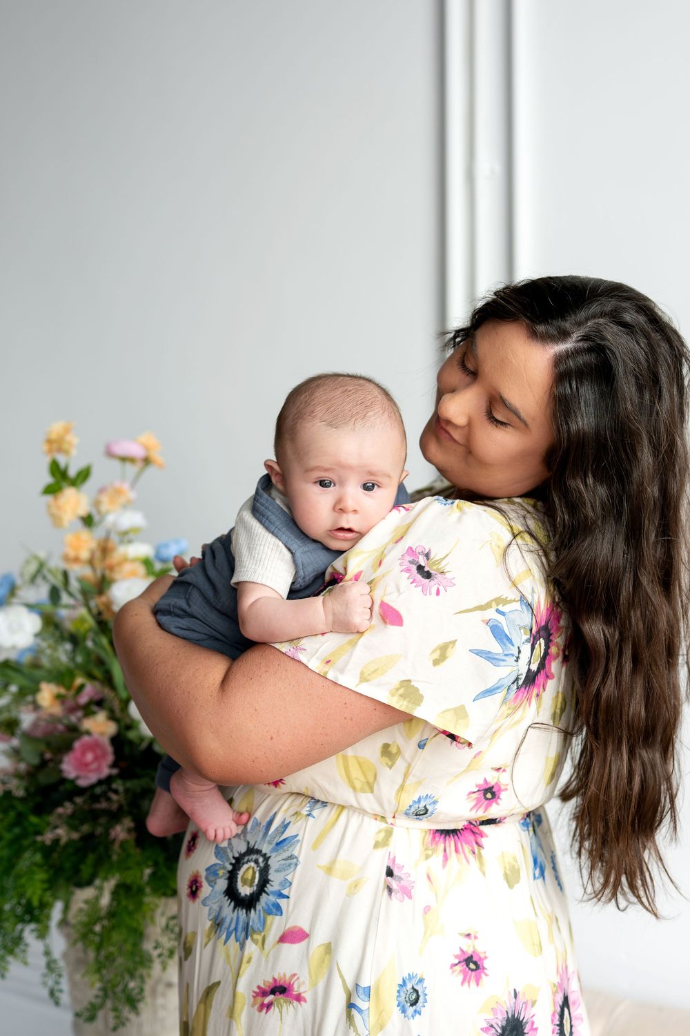Mother in floral dress tenderly holding alert newborn baby beside pastel flower arrangements indoors.