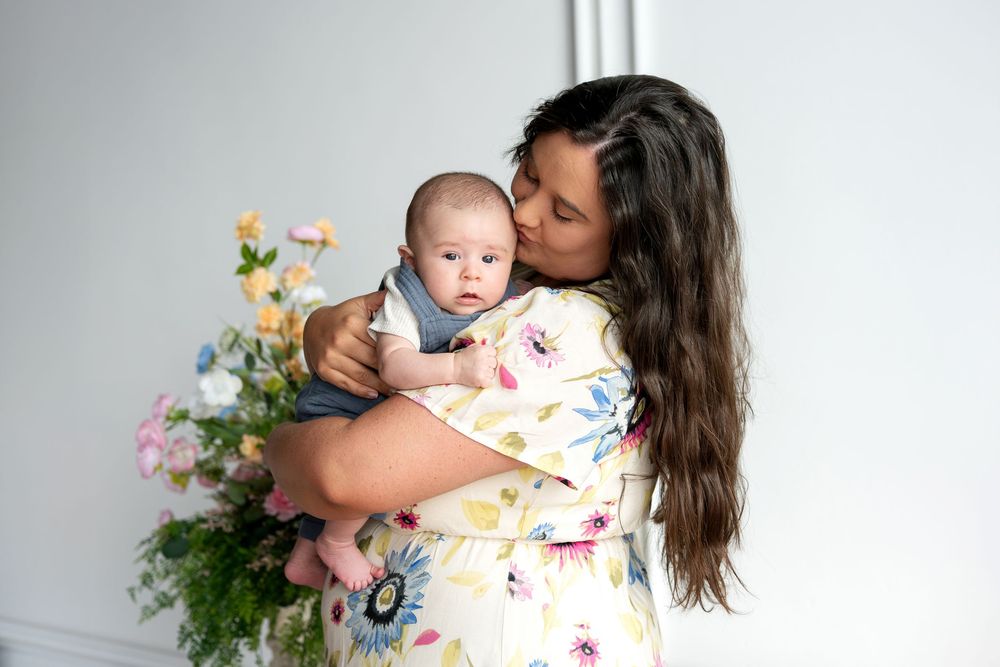 Mother in floral dress gently kissing newborn baby cheek beside colorful flower bouquet indoors.