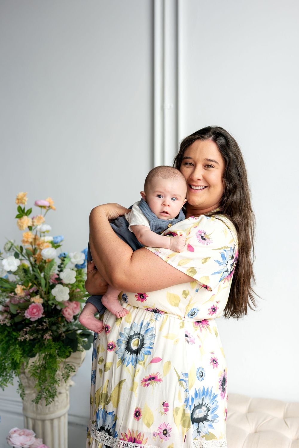 Smiling mother in floral dress holding wide-eyed newborn baby beside tall floral arrangement indoors.