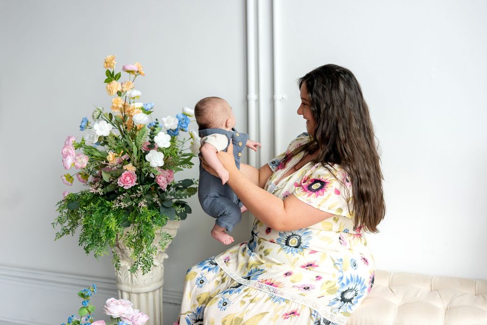 Mother in floral dress lifting newborn baby while seated beside tall elegant floral arrangement.