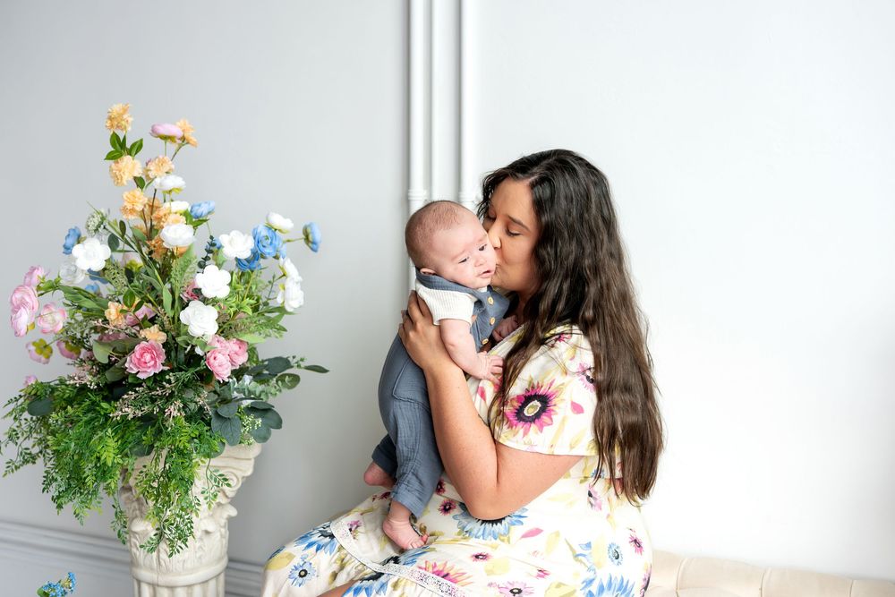 Mother in floral dress sitting and kissing newborn baby beside large pastel floral arrangement indoors.