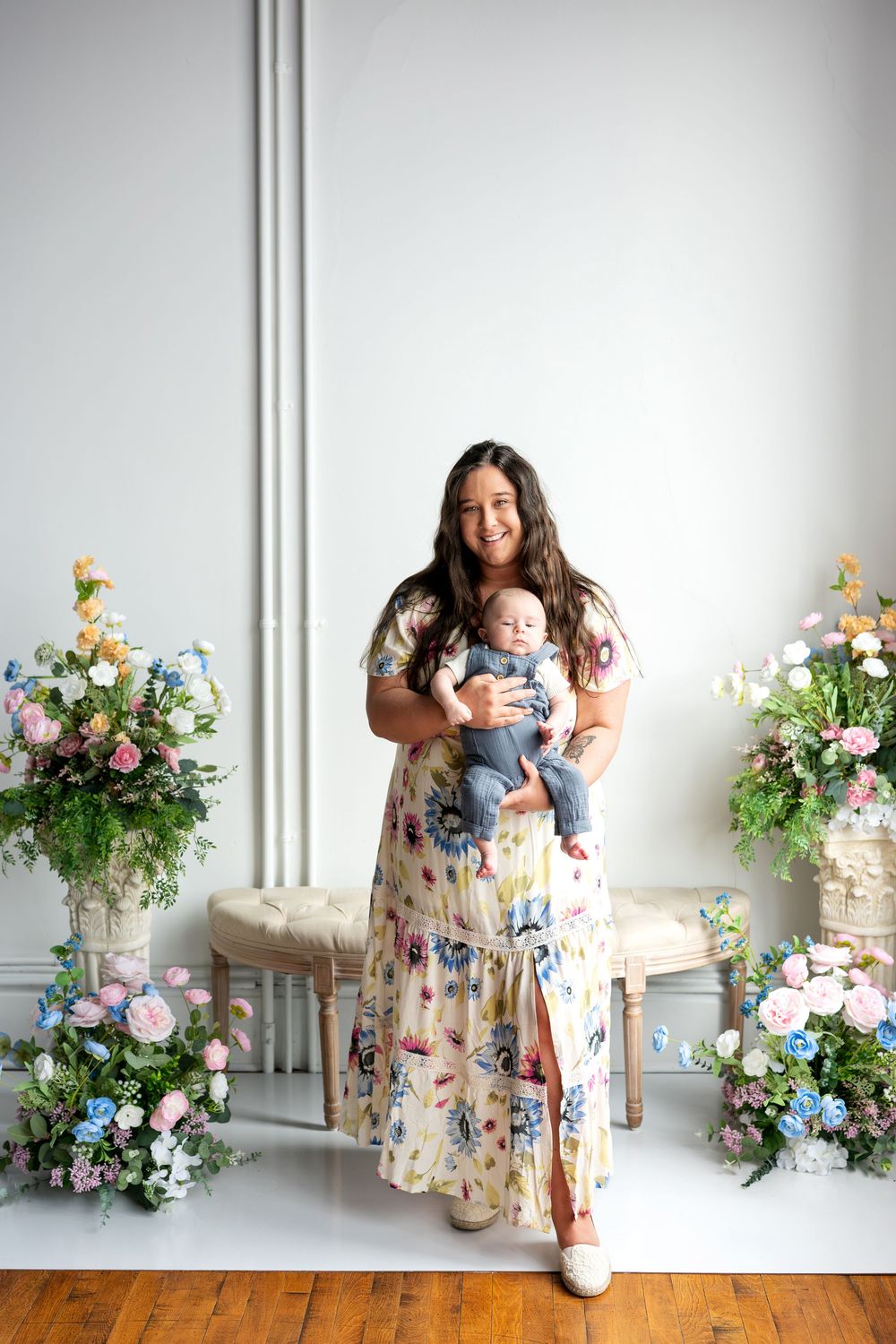 A smiling mother in a floral dress holds her baby boy in a studio with floral arrangements.