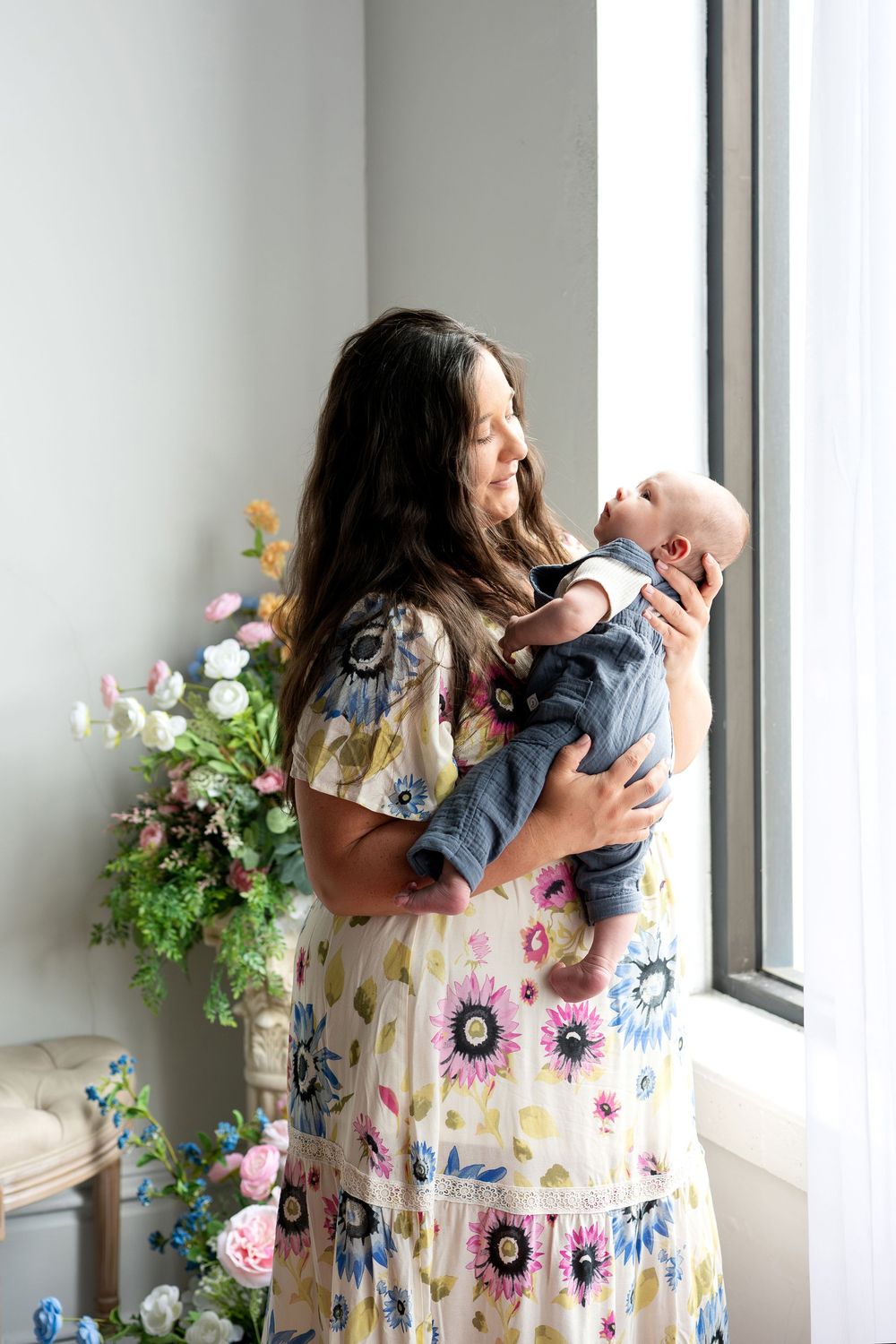 A woman in a floral maxi dress holds her newborn baby near a bright window with flowers.