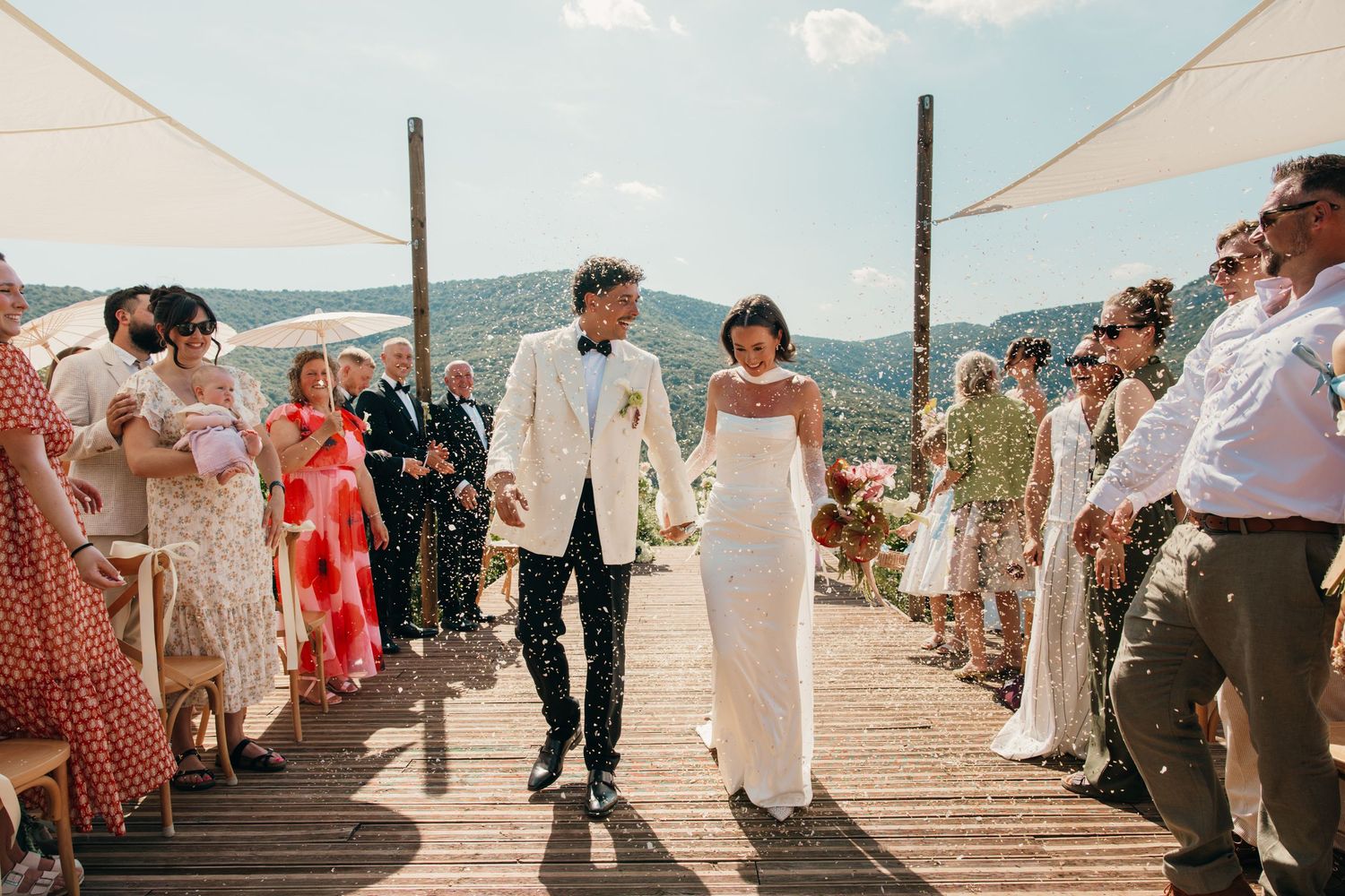 Bride in white gown and veil walks with groom in gray suit outside ivy-covered stone venue at golden hour.