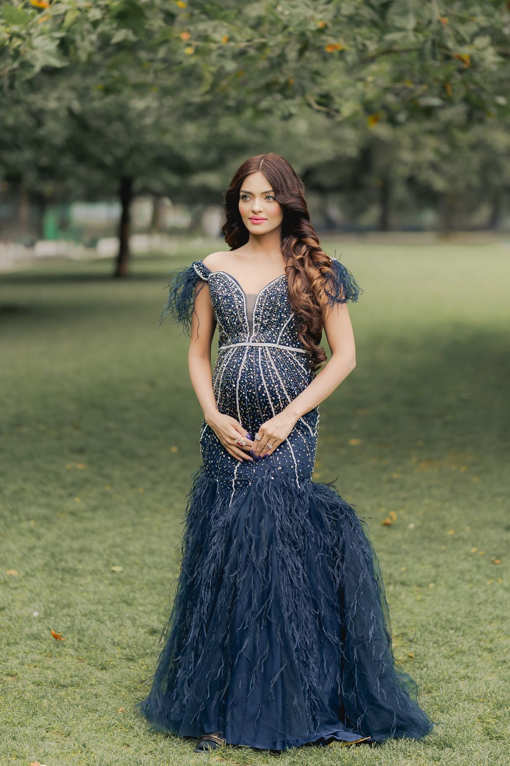 Pregnant woman in navy blue beaded mermaid gown with feather details poses outdoors in lush green park.