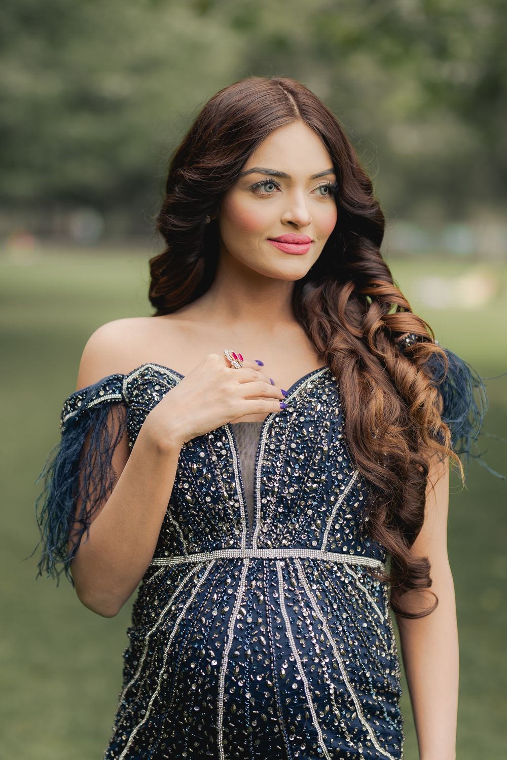 Woman in navy beaded off-shoulder gown with curly hair poses outdoors in a lush green park setting.