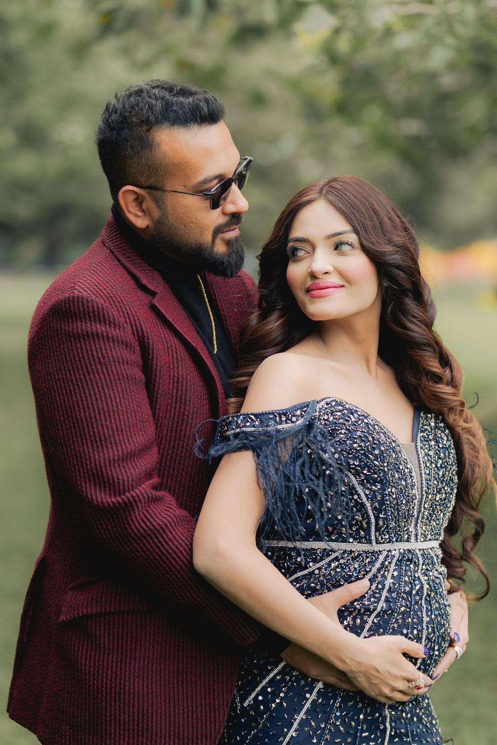 A couple poses outdoors, man in burgundy suit with sunglasses embracing woman in beaded navy off-shoulder gown.