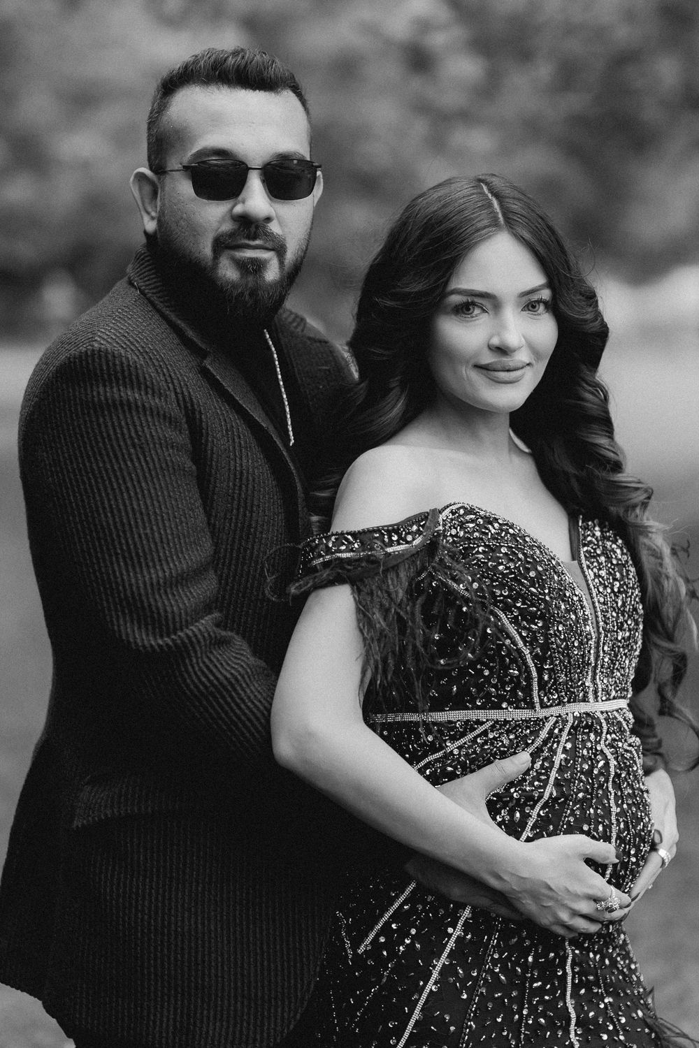 Black and white photo of an elegant couple posing outdoors, woman in beaded gown, man in suit with sunglasses.