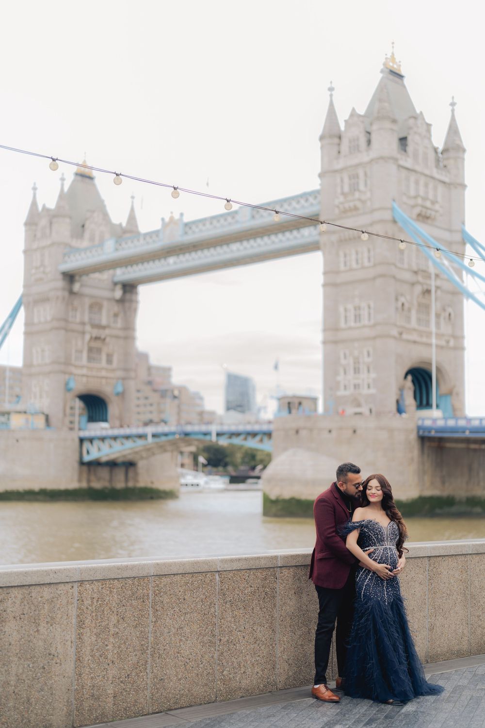 Couple posing romantically in front of Tower Bridge London, woman in navy gown and man in burgundy suit.