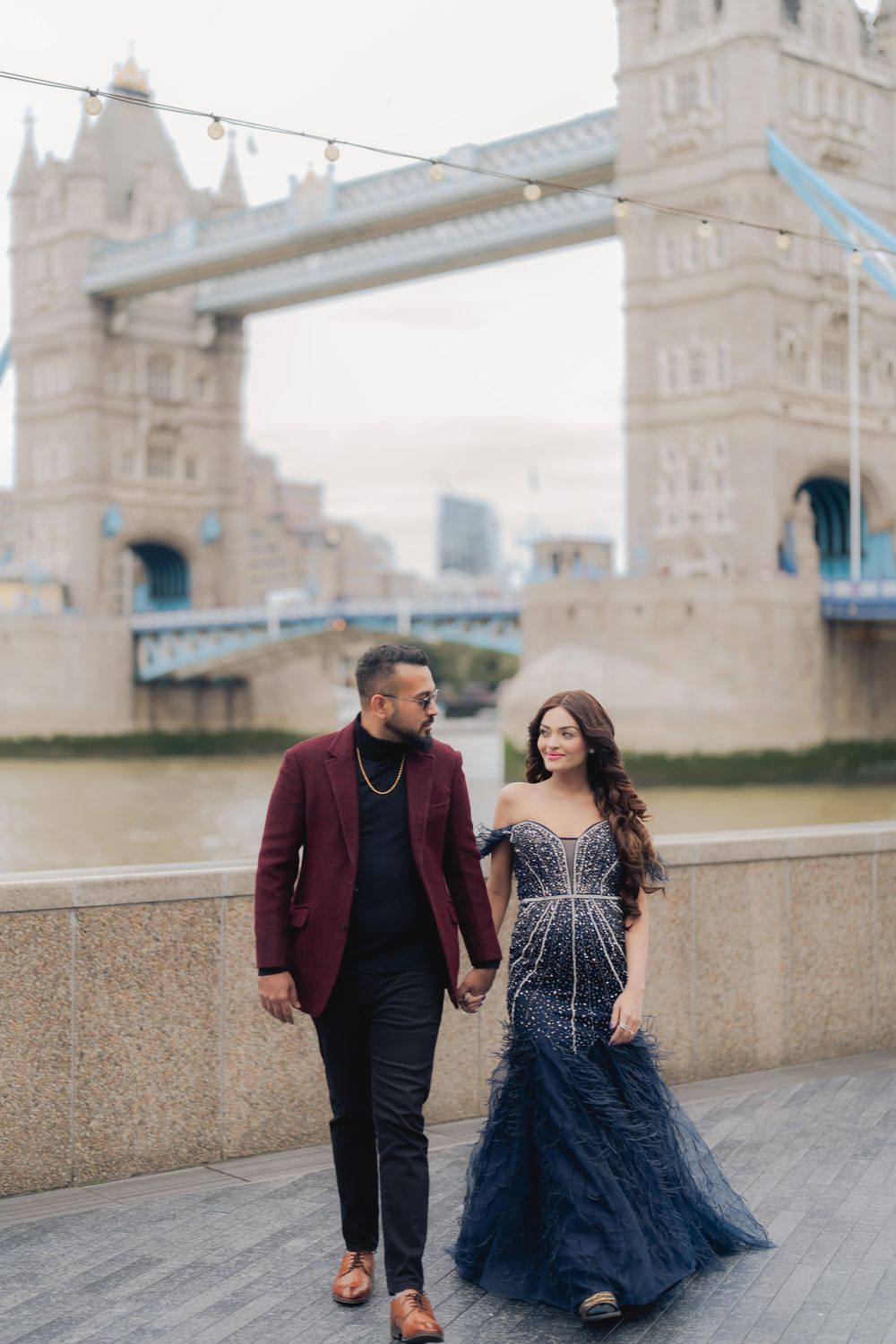 Couple in elegant attire holding hands near Tower Bridge London, man in burgundy blazer, woman in navy beaded gown.