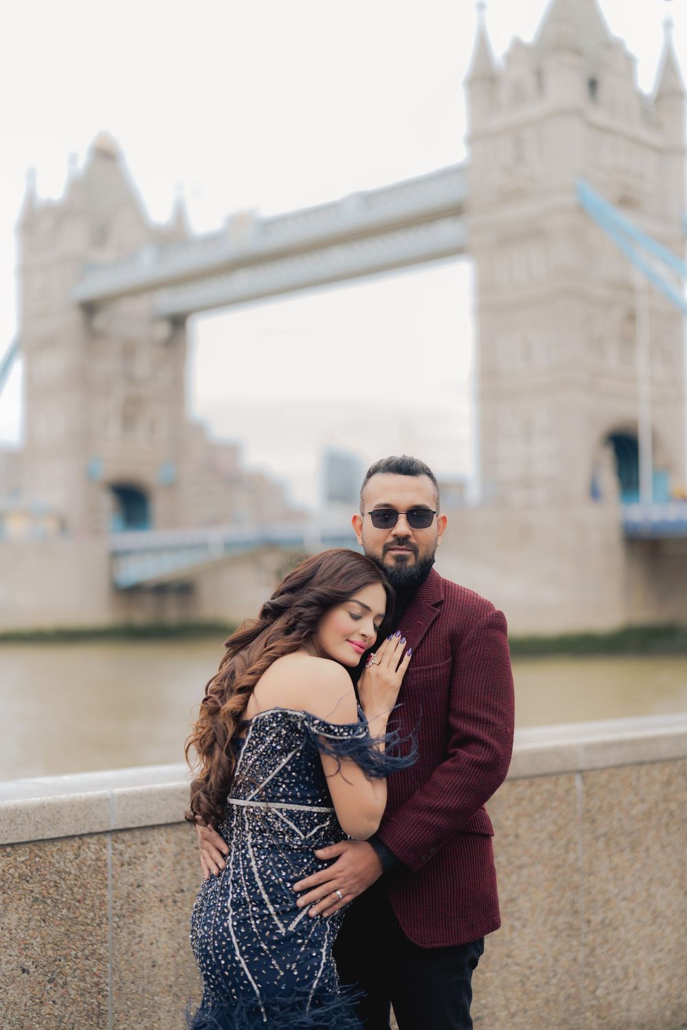 Couple embracing in front of Tower Bridge, London, woman in beaded gown, man in burgundy suit with sunglasses.