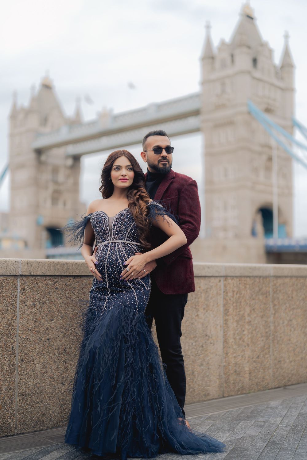 Elegant couple posing in formal attire before Tower Bridge London, woman in navy feathered gown, man in burgundy blazer.