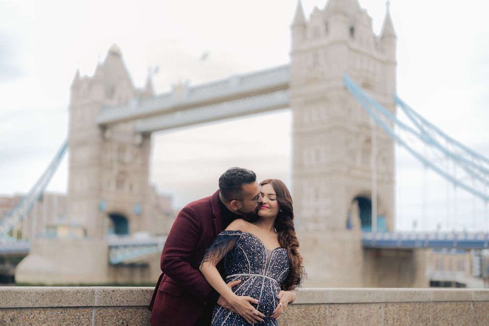 Couple embracing romantically in front of Tower Bridge in London, woman in blue gown, man in burgundy suit.
