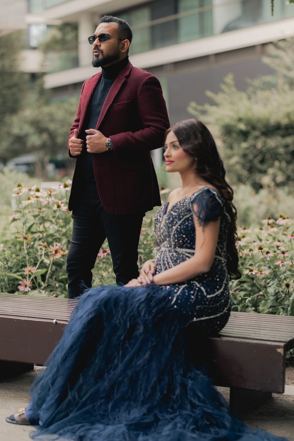 Elegant couple posing outdoors, man in burgundy blazer standing, woman seated in navy beaded gown.