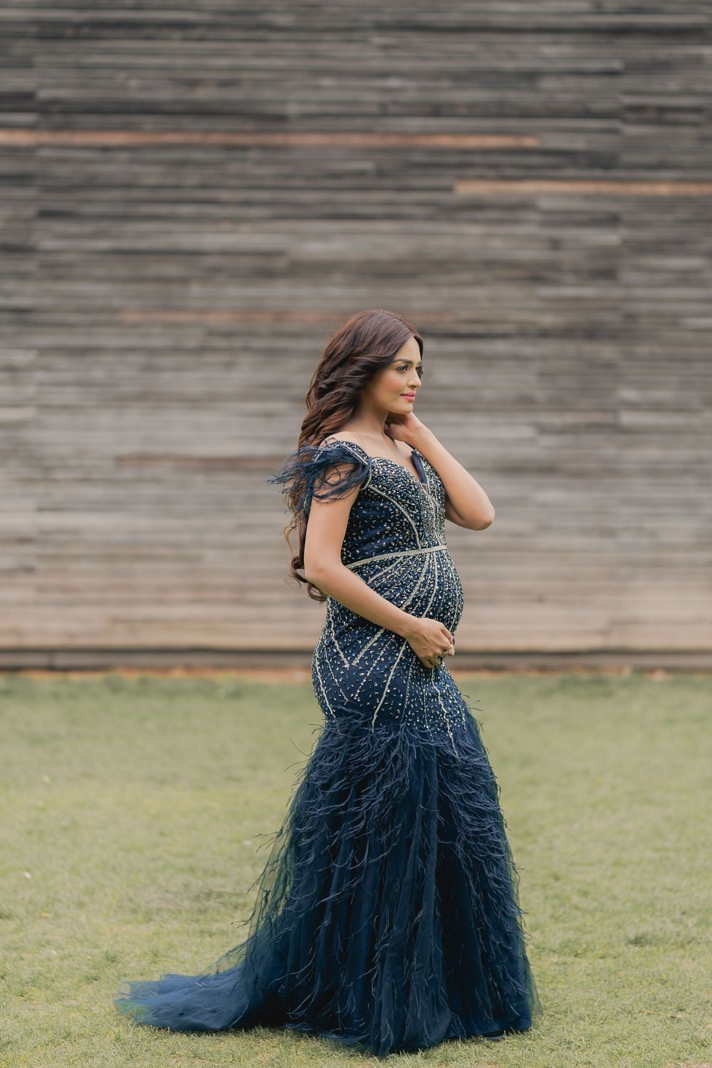 Pregnant woman in navy blue beaded mermaid gown with feather trim posing outdoors against wooden wall.