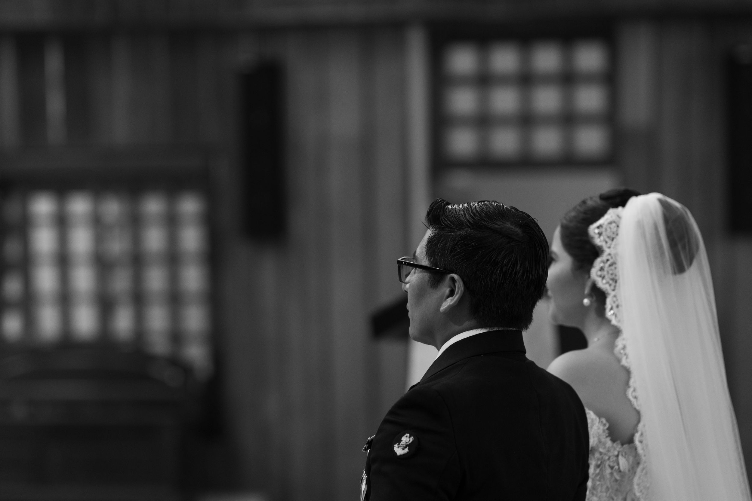Black and white wedding photo of bride in lace veil and groom in glasses standing at the altar.