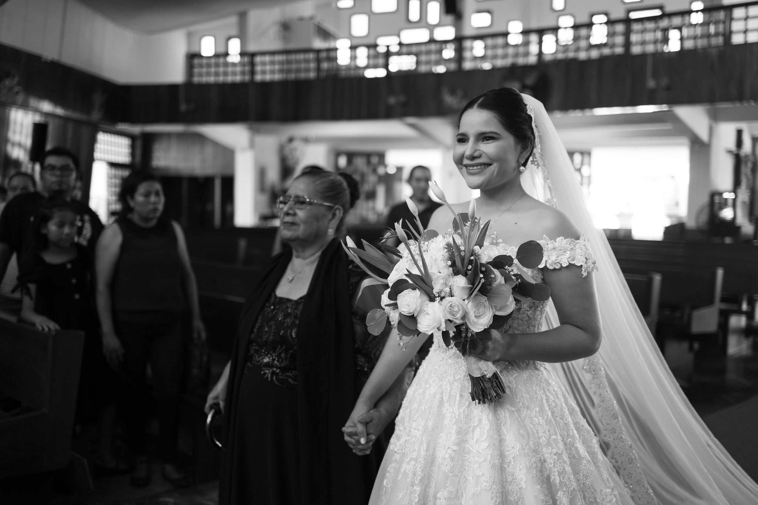 Black and white photo of a smiling bride in a lace gown holding a bouquet, walking down the aisle with guests.