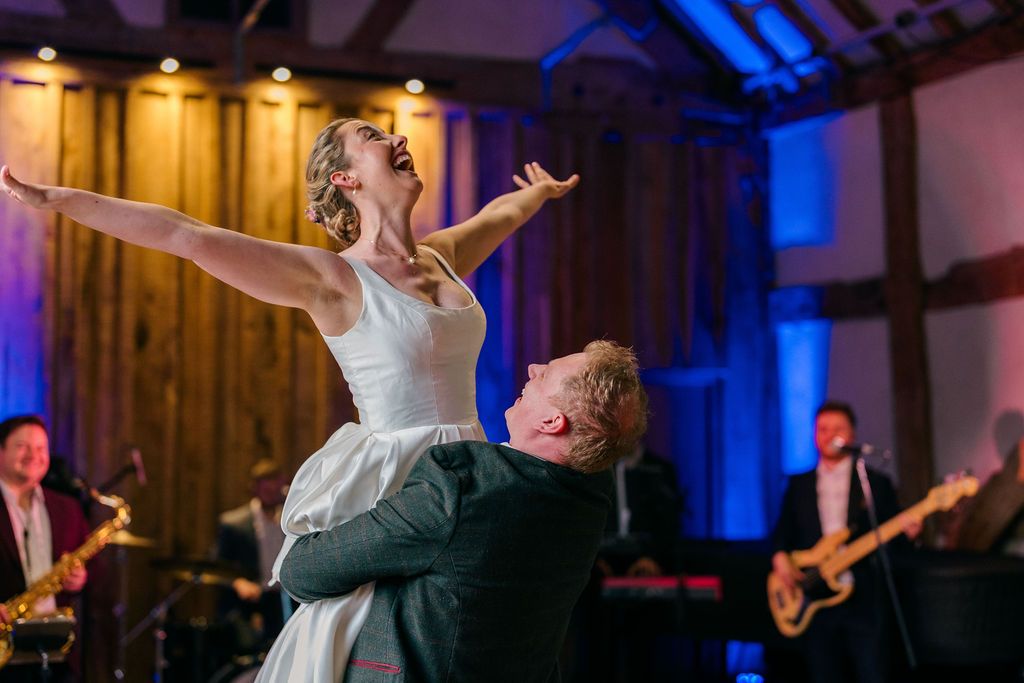 Bride lifted by groom during first dance at wedding reception with live band performing in background.
