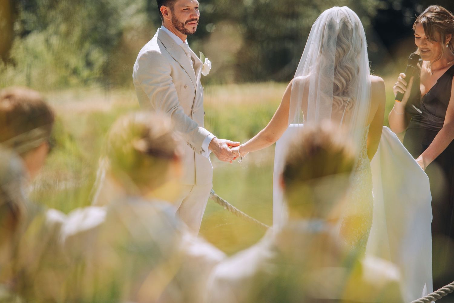 Outdoor wedding ceremony with smiling groom in tan suit holding hands with bride in white veil and dress.