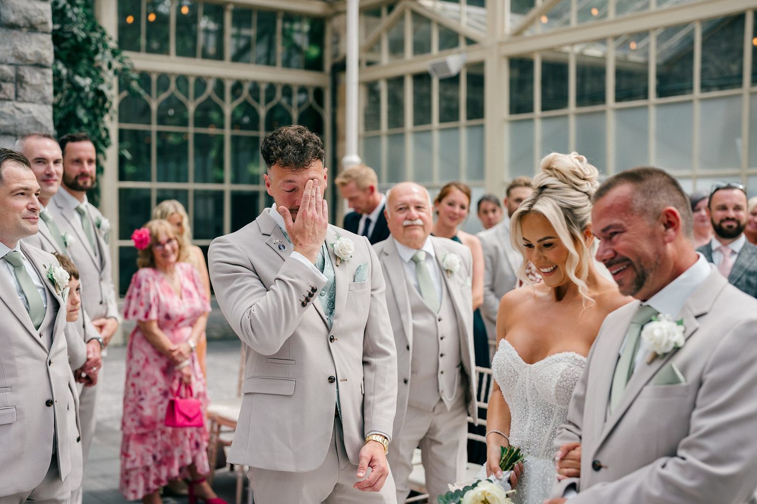Emotional groom crying as bride walks down the aisle at elegant greenhouse wedding ceremony with guests watching.