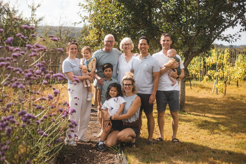 Multi-generational family posing together outdoors near vineyard and purple wildflowers on a sunny day.