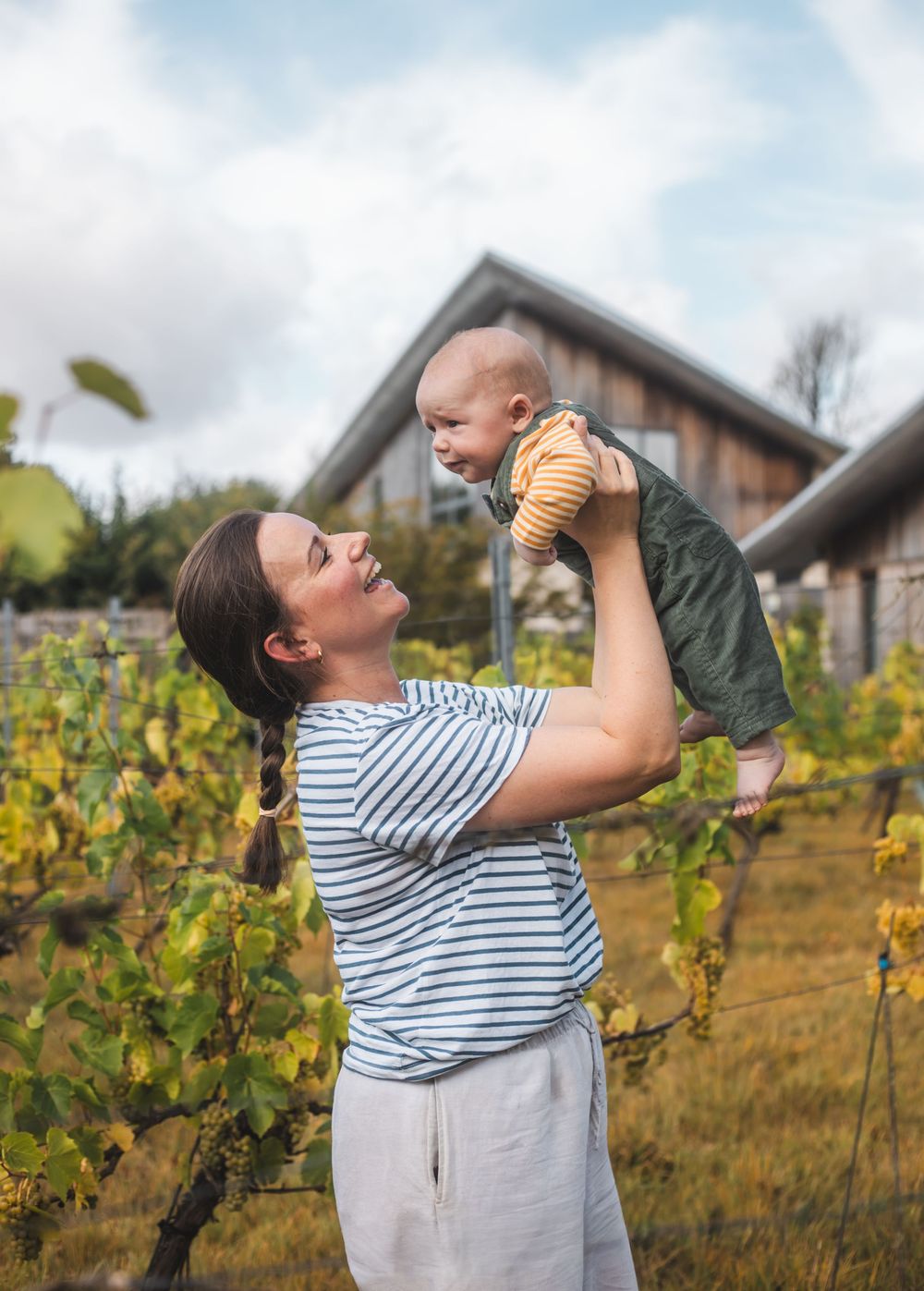 Mother joyfully lifts smiling baby in vineyard with rustic barn backdrop on cloudy day.