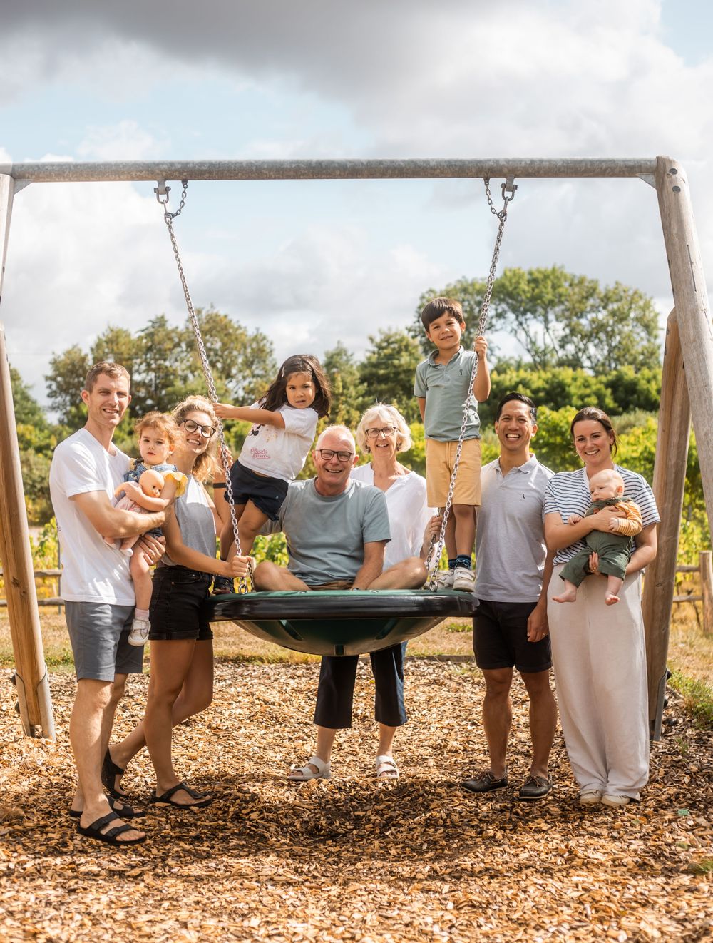 Multi-generational family posing together at outdoor playground with nest swing surrounded by greenery.