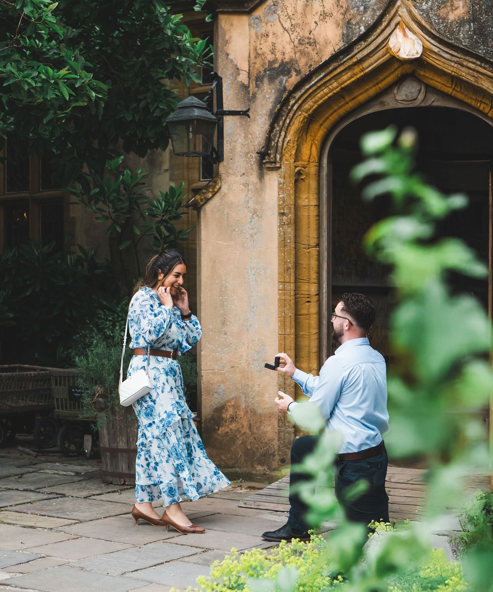 Man proposing to woman in floral dress outside a historic stone archway building.