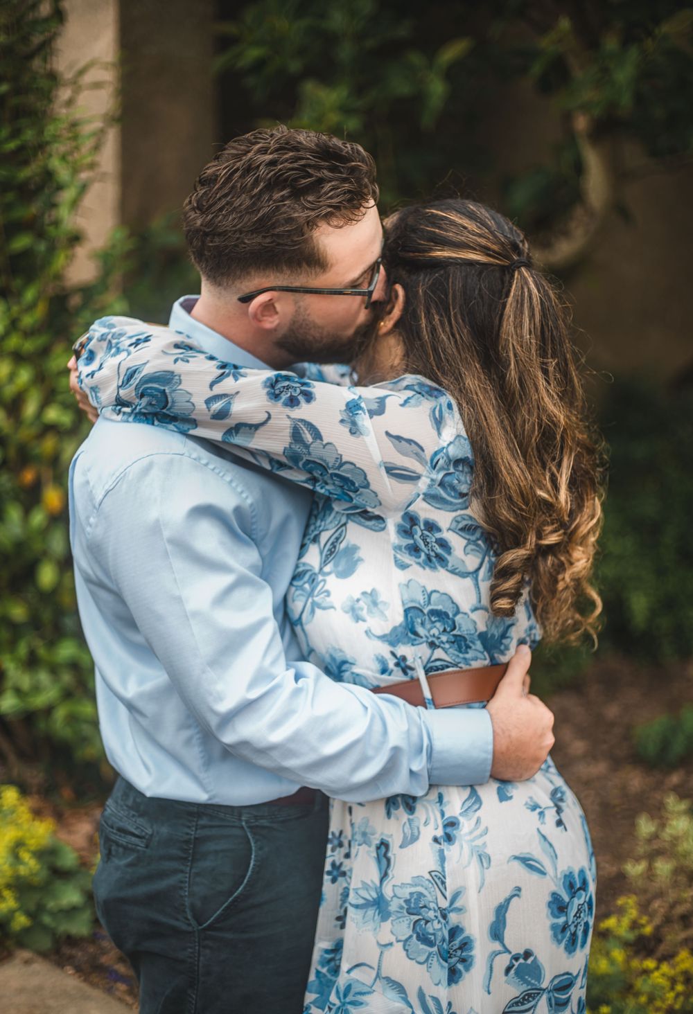A couple shares a romantic kiss outdoors, with the woman wearing a white and blue floral dress.
