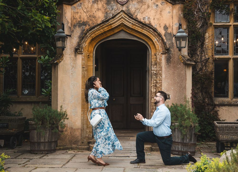 Man proposes on one knee to surprised woman in floral dress outside a historic stone building.
