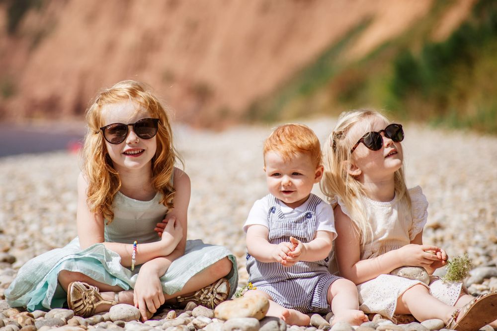 Three young children sitting on a pebble beach, two girls in sunglasses flanking a smiling baby in summer outfits.