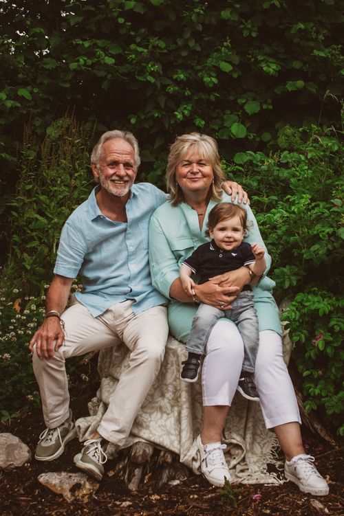 Grandparents sitting on a rock with their toddler grandchild in a lush garden.