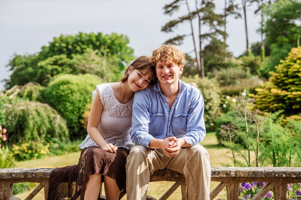 A smiling couple sitting together on a garden bench surrounded by lush greenery on a cloudy day.