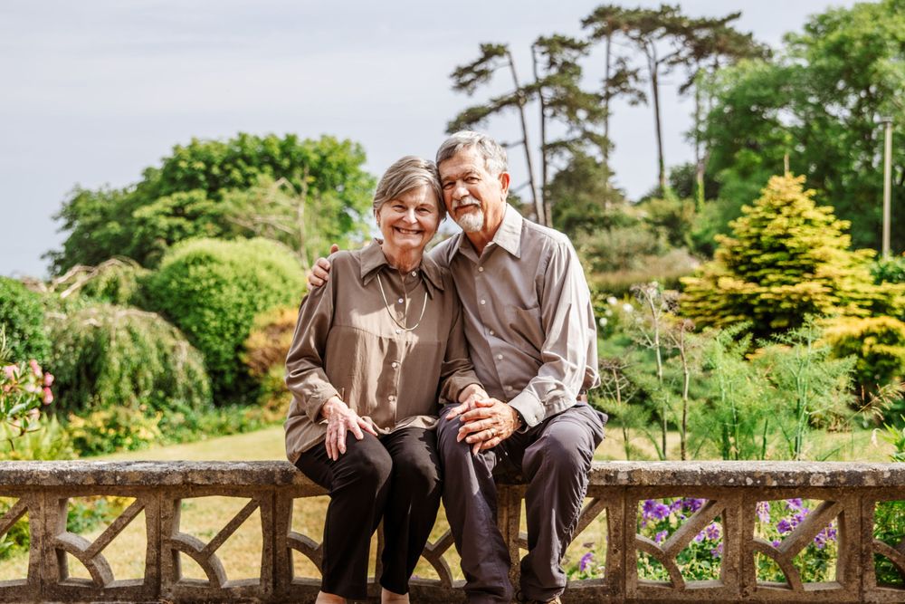 Happy elderly couple sitting together on a stone bench in a lush garden, smiling warmly on a cloudy day.